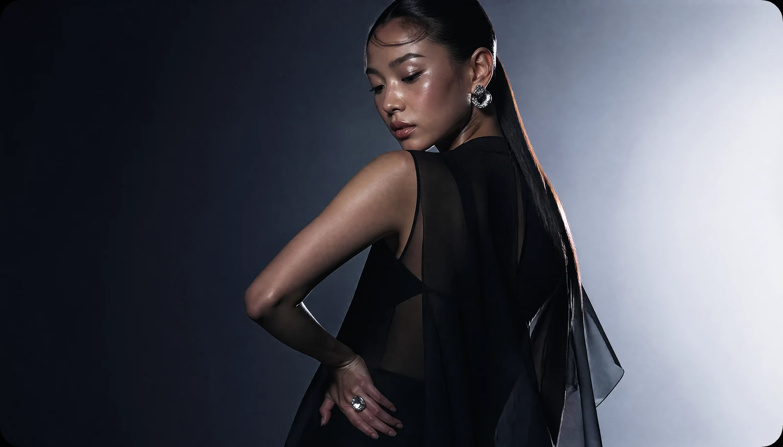 Luxury fashion editorial with a black satin cape, sculptural pose, silver jewelry, and a cool studio gradient.